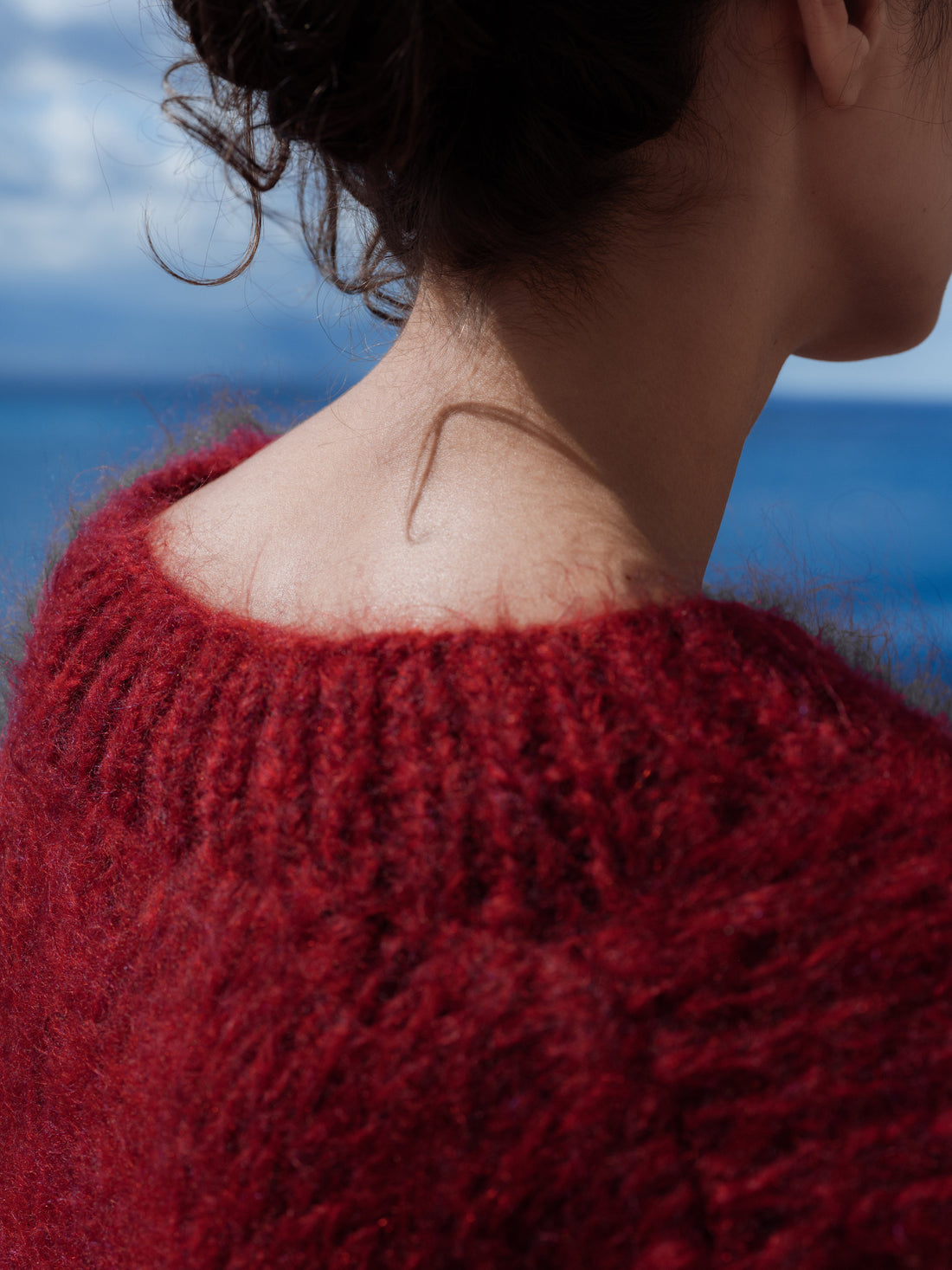 A woman wearing a signature Henry’s Pick made to order baby-mohair maxi sweater, shown is the part where the wool cherishes her neck. in the background the blue mediterrean sea.