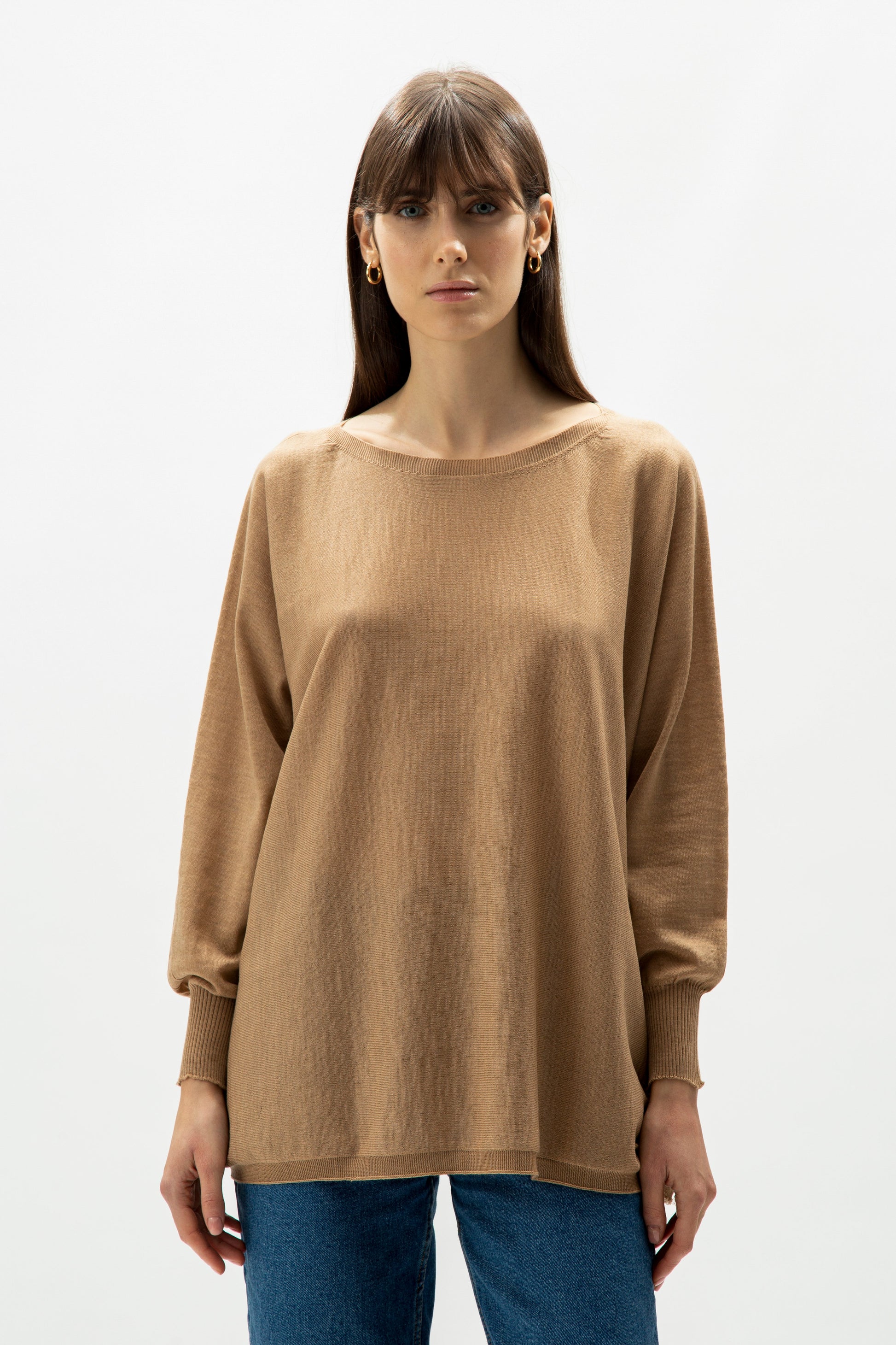 Woman wearing a loose camel knit sweater and blue jeans, minimalist studio background