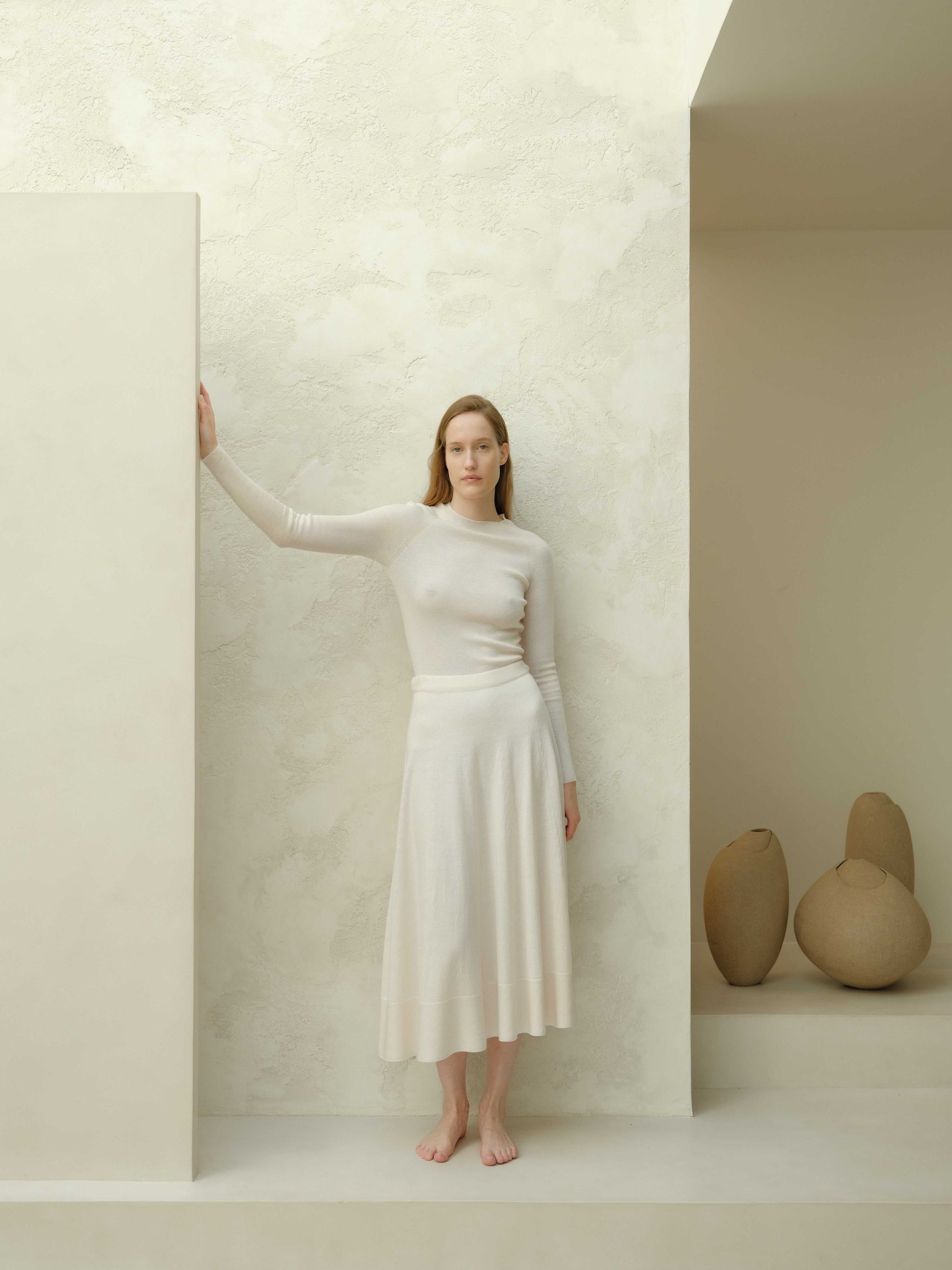 Full-body view of ivory slim fit ribbed crewneck in extra fine merino wool, styled with matching skirt. Seamless knitwear in a serene setting.