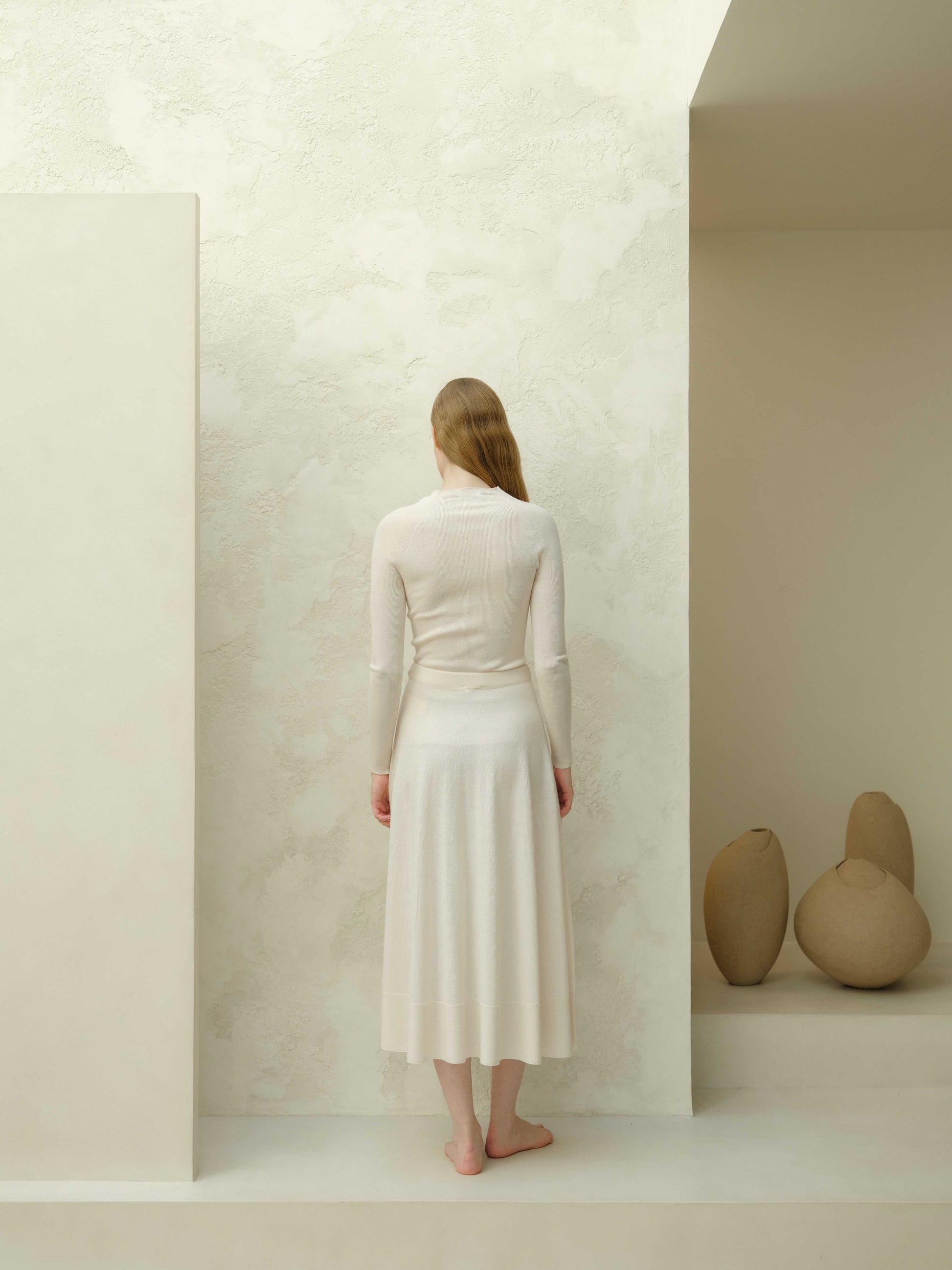 Rear view of ivory slim fit ribbed crewneck in extra fine merino wool, paired with matching skirt. Minimalist knitwear with a soft drape.
