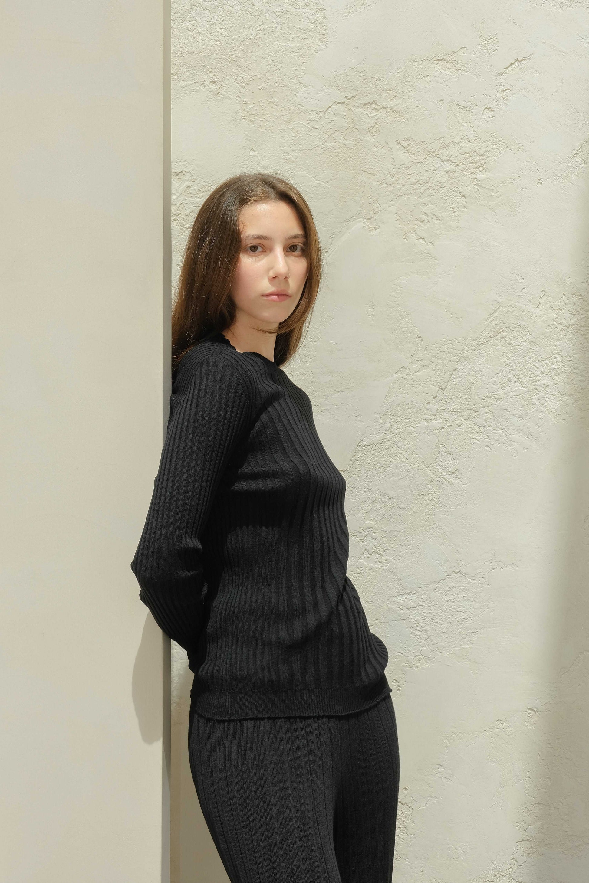 Extra Fine Merino Wool Ribbed Crew Neck 3D-Knit Sweater in black, slim-fit and seamless, modeled against a textured light wall.