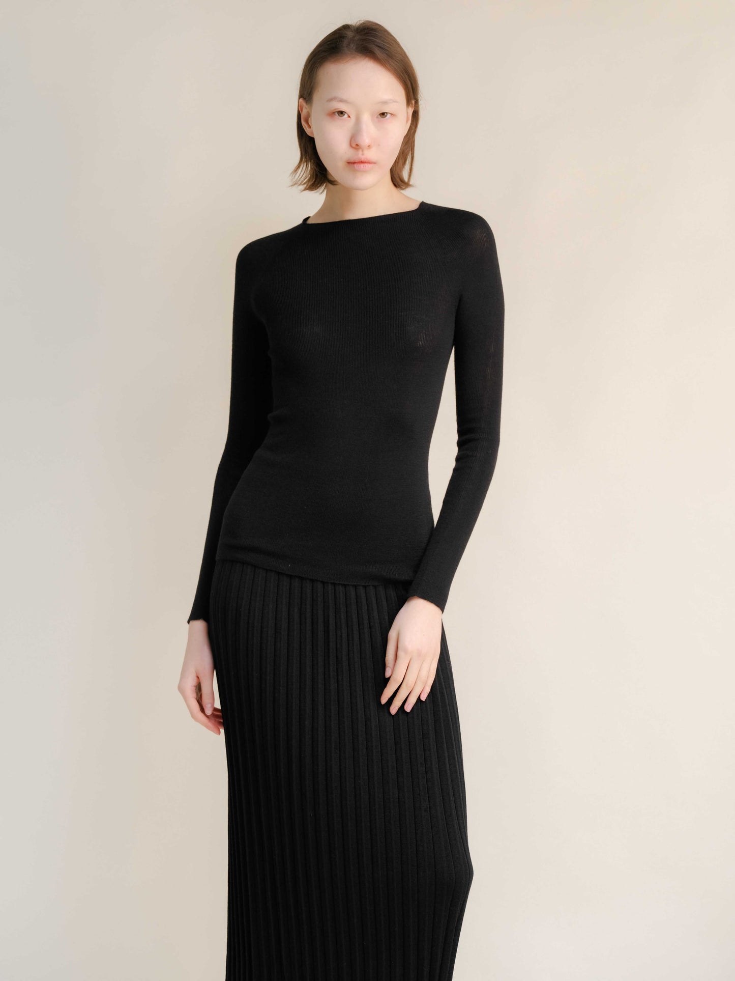 Black slim fit ribbed crewneck in extra fine merino wool, styled with pleated skirt. Seamless knitwear for versatile and elegant layering.