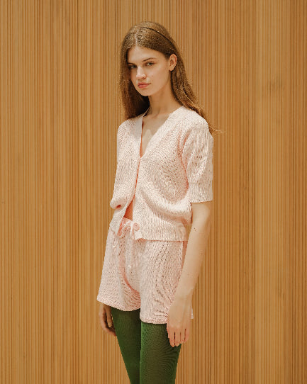 Woman wearing a light pink cardigan and matching pants against a wooden paneled wall.