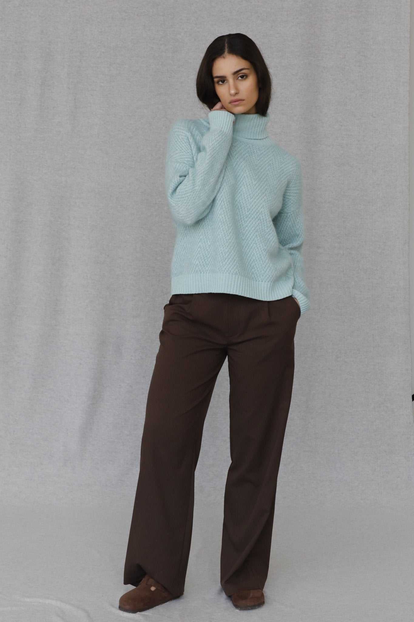 Penelope Chevron Wool Turtleneck in soft blue merino wool, paired with brown trousers. Cozy chevron knit for cold-weather wear.