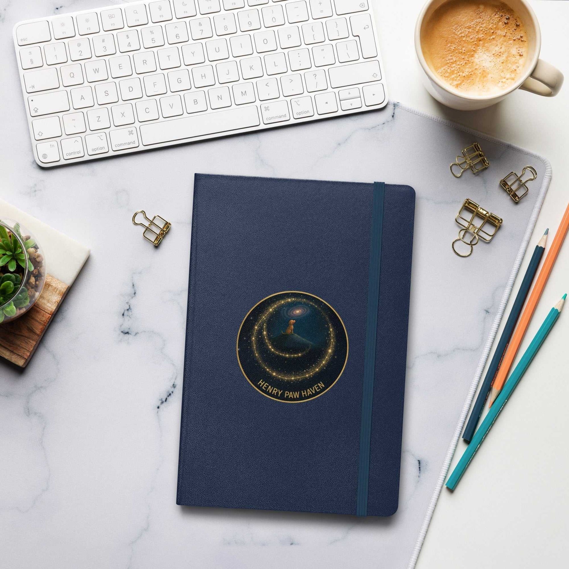 Navy blue Hardcover Bound Notebook 5.5" × 8.5" with elastic band and HenryPawHaven logo, placed on a workspace with keyboard and coffee.
