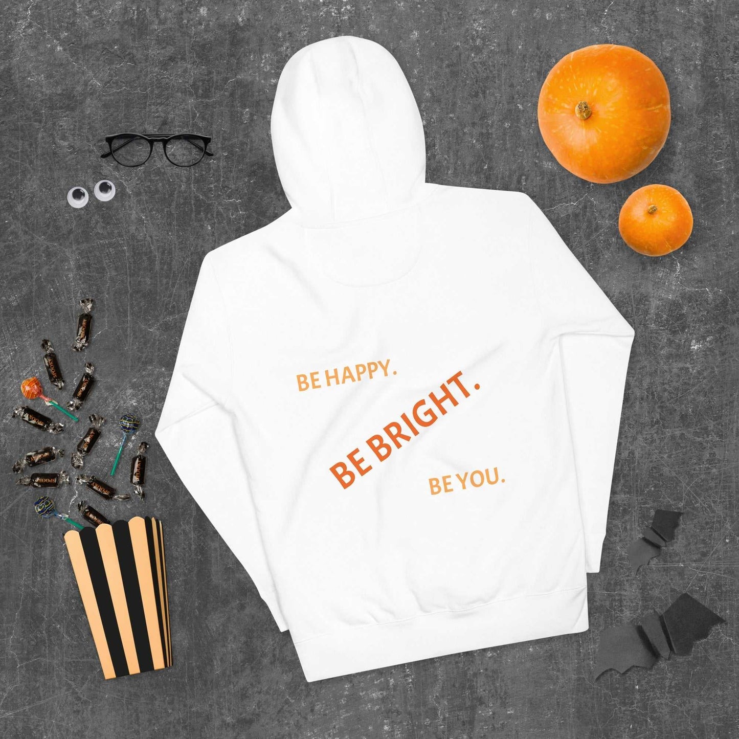 White unisex hoodie laid flat with bold orange motivational text on back, styled with Halloween decor for festive comfort.