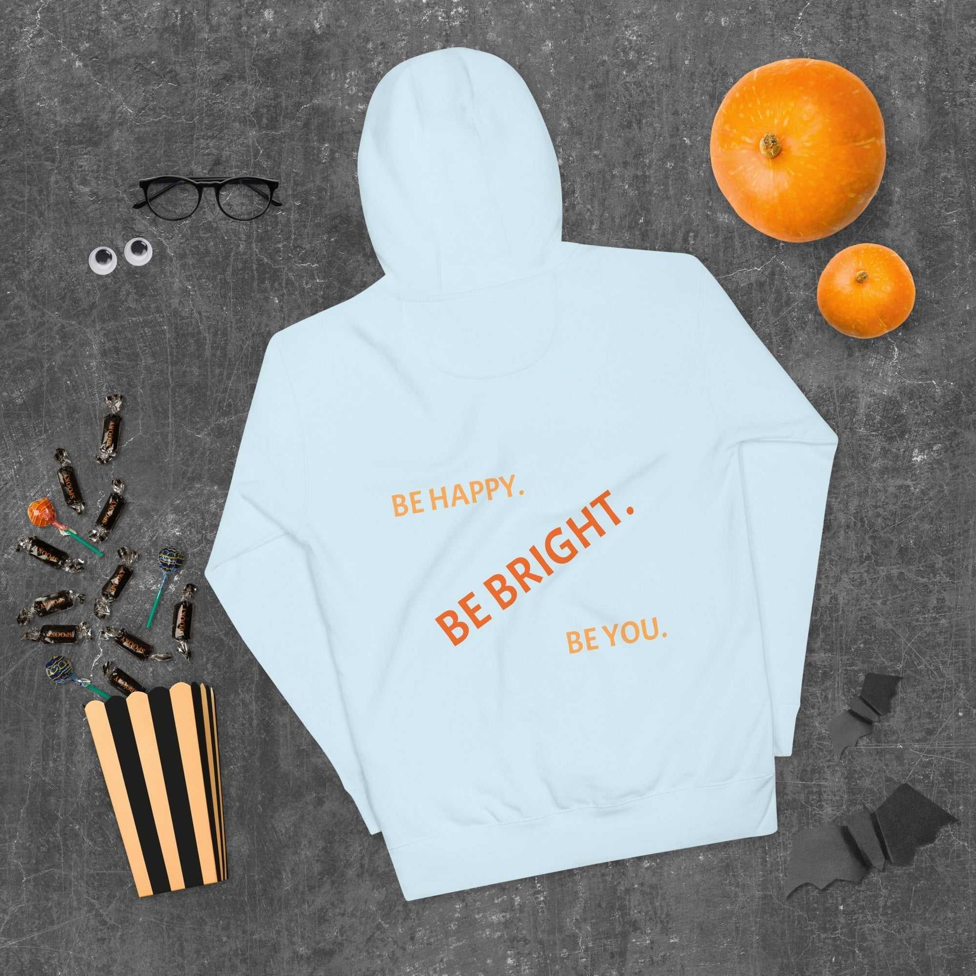 Light blue unisex hoodie laid flat with 'Be Happy. Be Bright. Be You.' text, surrounded by Halloween items for festive comfort.
