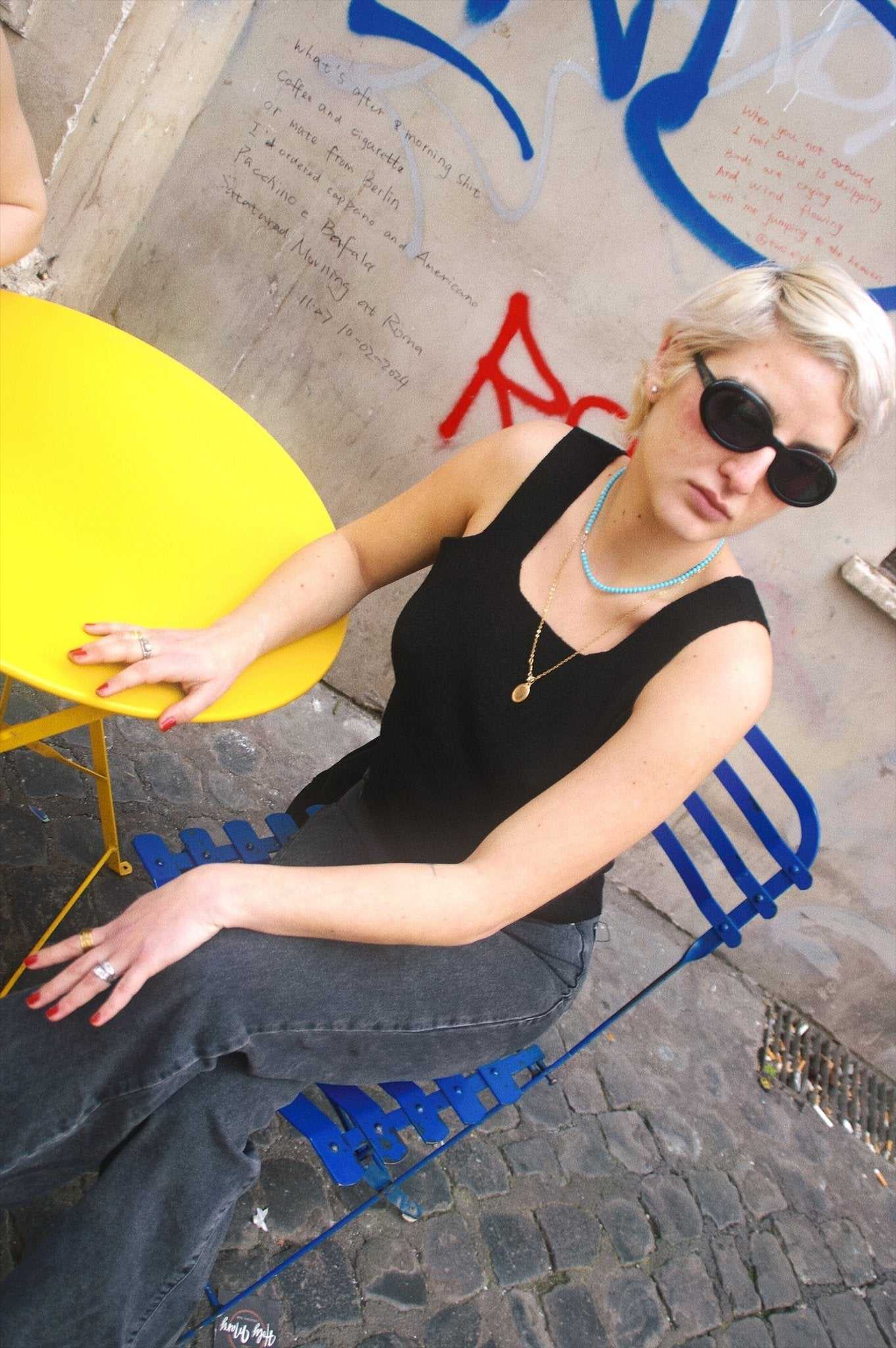 Margaret Tank Top - Sustainable Lyocell Tank Top in black styled with dark jeans and layered necklaces, worn by woman seated at a graffiti-covered café wall.