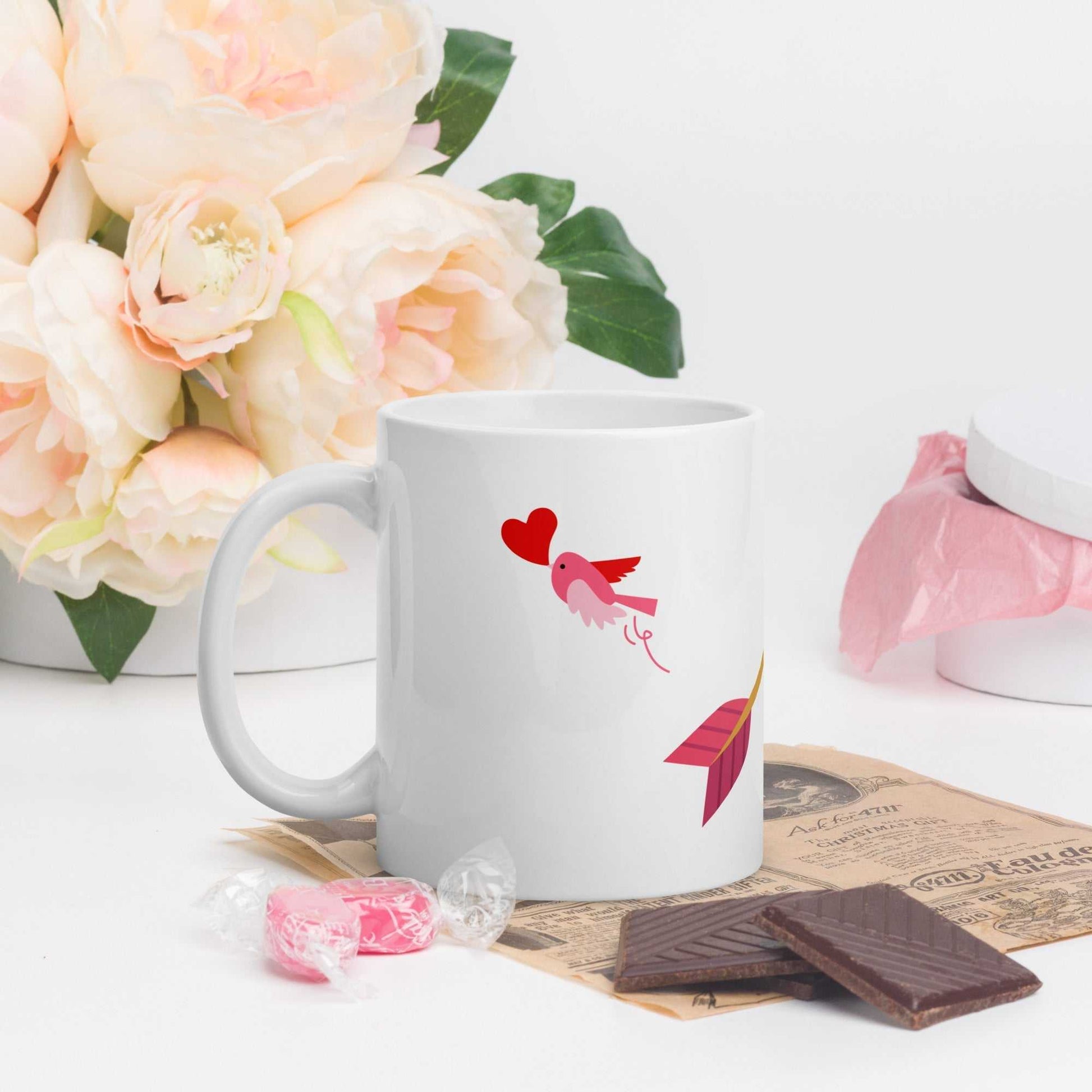 Valentine mug gift with pink bird and heart arrow design, styled with flowers and chocolates for a romantic setting.