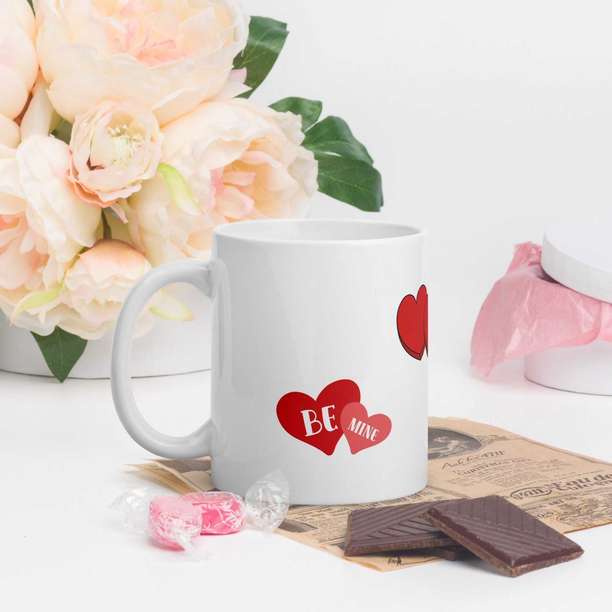 11 oz ceramic Valentine mug with red heart and 'Be Mine' design, placed beside flowers and chocolates, ideal for romantic gifting.