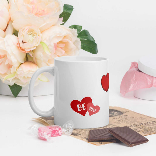 11 oz ceramic Valentine mug with red heart and 'Be Mine' design, placed beside flowers and chocolates, ideal for romantic gifting.