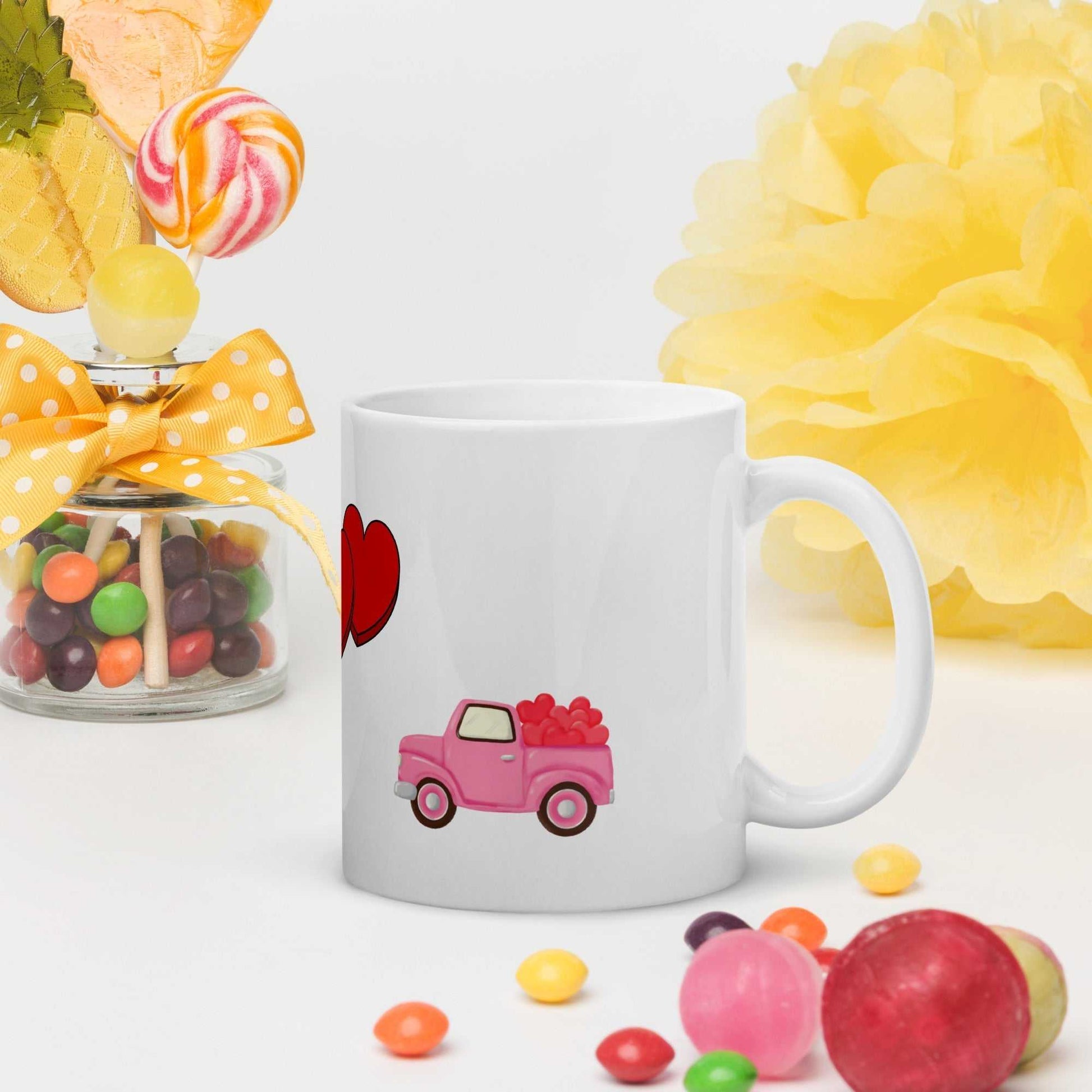 White ceramic Valentine mug with pink truck and heart design, styled with candy and yellow decor for festive gifting.
